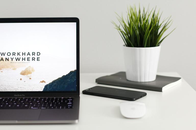 A dark grey laptop on a white desk with a black smartphone and green plant to its right