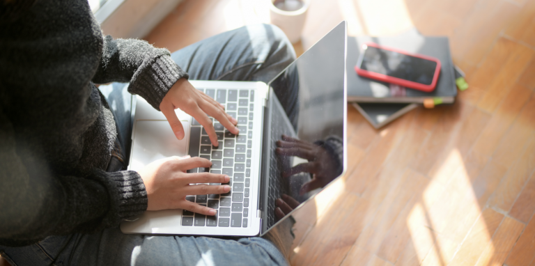 A person sat cross-legged typing on a silver laptop