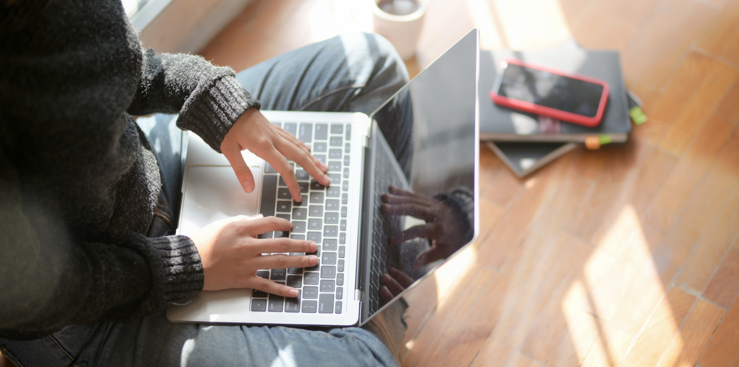A person sat cross-legged typing on a silver laptop