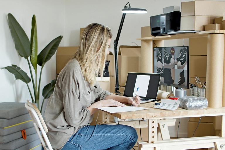 Woman sat at a desk working on a laptop surrounded by boxes of products setting up a Shopify store.